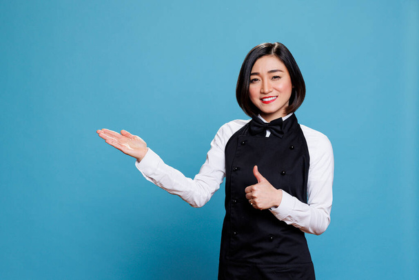 Gai asiatique femme réceptionniste montrant pouce vers le haut et pointant vers le côté avec portrait à la main. Serveuse montrant un geste de satisfaction et montrant la bonne direction tout en regardant la caméra - Photo, image