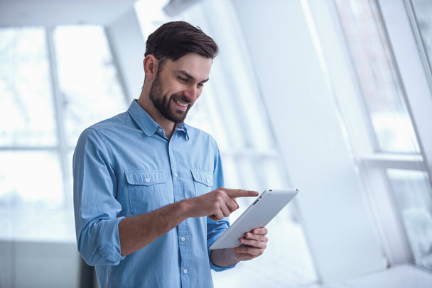 Bello uomo d'affari sta utilizzando un tablet digitale e sorridente mentre in piedi in ufficio
 - Foto, immagini