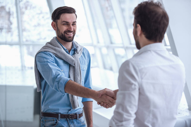 Два привлекательных бизнесмена пожимают руки и улыбаются стоя на посту
 - Фото, изображение