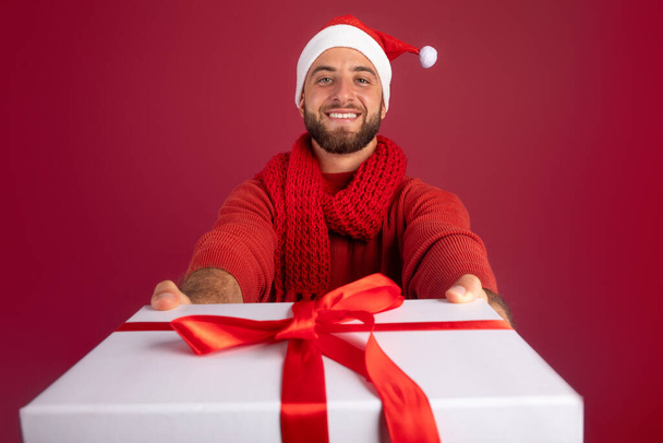 Happy millennial caucasian guy with beard in Santa hat give box of gift isolated on burgundy studio background, close up. Christmas, New Year holidays, ad and offer, present idea - Valokuva, kuva