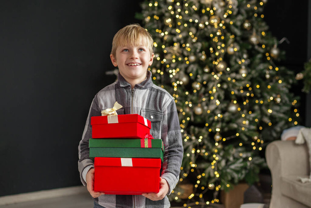 Elinde hediye kutuları olan bir çocuk Noel ağacı arka planında. Kavram Yeni Yıl, Mutlu Noeller, tatil, kış, çocukluk. - Fotoğraf, Görsel