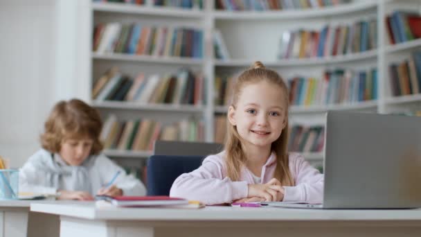 Концепция образования. Милая маленькая школьница жестикулирует большой палец вверх улыбаясь в камеру, обучение на ноутбуке с детьми в современном классе в школе, замедленная съемка, свободное пространство - Кадры, видео