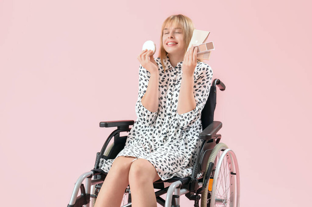 Ευτυχισμένη νεαρή γυναίκα σε αναπηρική καρέκλα με διακοσμητικά καλλυντικά σε ροζ φόντο - Φωτογραφία, εικόνα