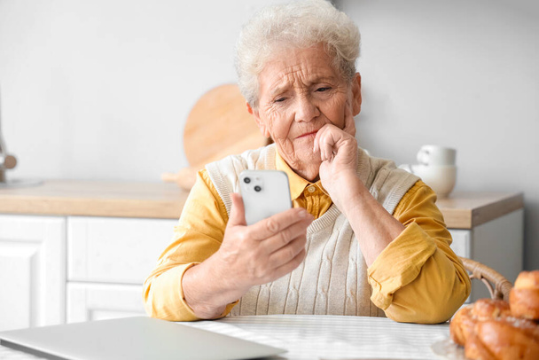 Femme âgée réfléchie utilisant le téléphone mobile dans la cuisine - Photo, image