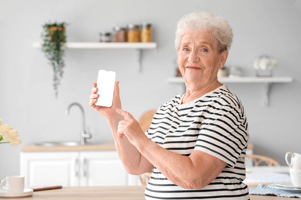 Mujer mayor señalando el teléfono móvil en la cocina - Foto, Imagen
