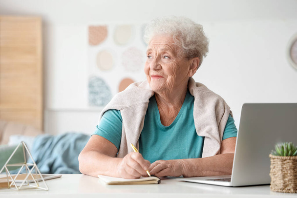 Seniorin mit Laptop schreibt zu Hause in Notizbuch - Foto, Bild