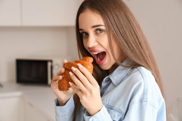 Gyönyörű fiatal nő eszik ízletes croissant a konyhában - Fotó, kép