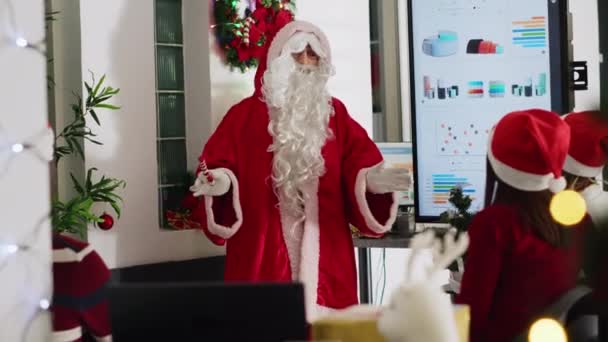 Supervisor vestido como Santa Claus durante la temporada navideña celebrando una reunión de equipo, mostrando los datos de la compañía en pantalla digital. Líder de equipo en el lugar de trabajo decorado de Navidad hablando con los empleados - Metraje, vídeo