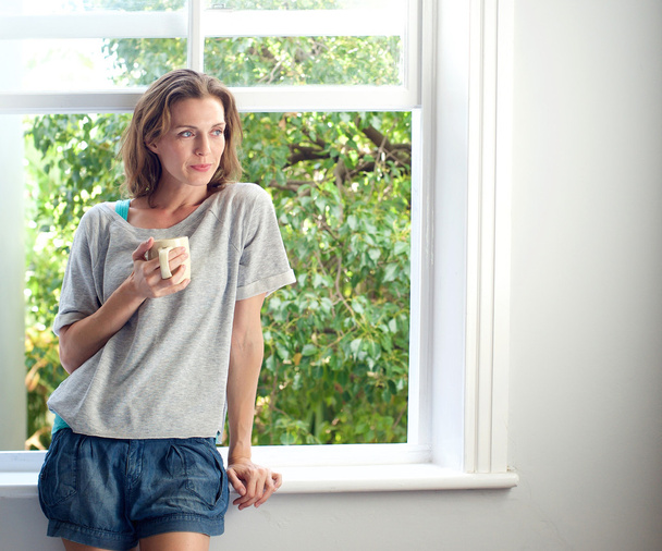 Mujer de pie junto a la ventana en casa con una taza de café
 - Foto, Imagen