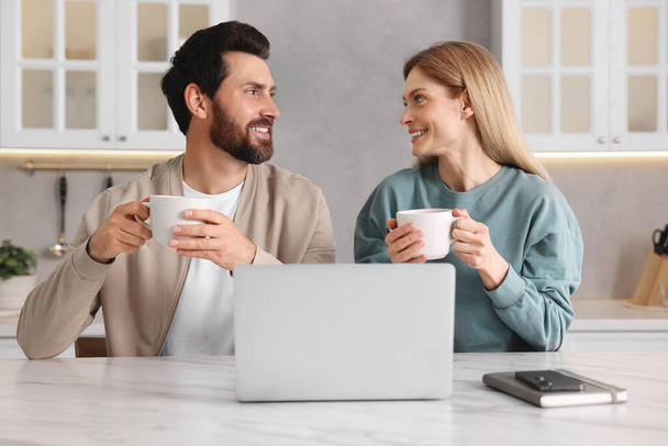 Happy couple with laptop and cups of drink at white table in kitchen - Фото, зображення