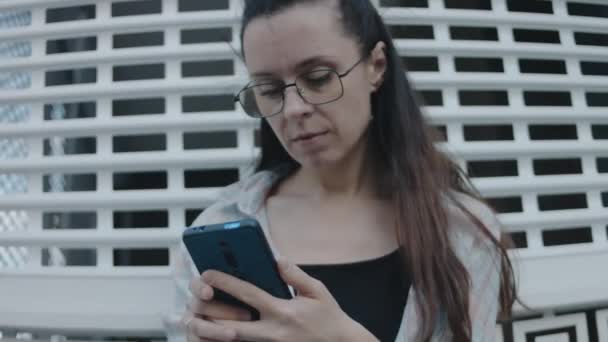 近代的な建物の近くに一人で立っている眼鏡を持つ若い女性,携帯電話を使用して,画面を見たり,誰かに話したり,アプリチャットでメッセージを送信したり, - 映像、動画