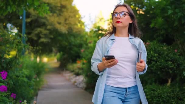 Mujer feliz con laptop y smartphone en sus manos caminando por el floreciente jardín o calle verde después del día de trabajo, cámara lenta. - Metraje, vídeo