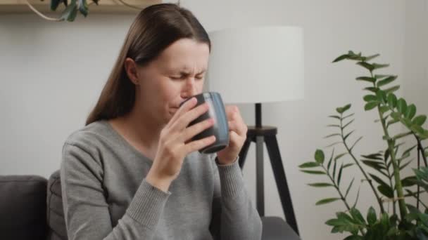 Sick unhappy young caucasian woman touch neck, have sore throat, holding mug, drink warm water, have a fever, flu in weakness, sitting alone on sofa at home. Health care on virus seasonal - Filmagem, Vídeo