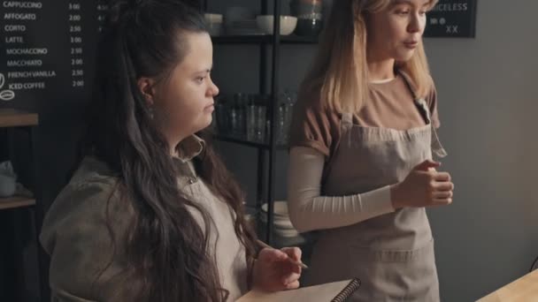 Közepes felvétel fiatal kaukázusi nőről, aki a pult mögött áll a modern kávézóban, és megmondja az új fogyatékos kollégájának, hogyan dolgozzon a megrendelésekkel. - Felvétel, videó