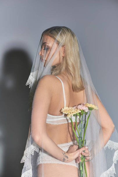 attrayant sexy femme en lingerie blanche avec voile tenant des fleurs derrière son dos sur fond gris - Photo, image