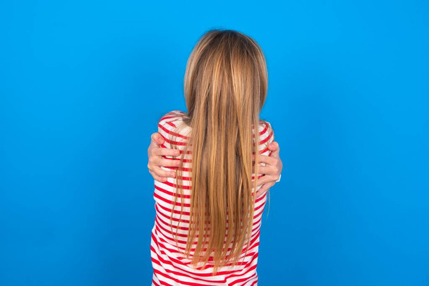 красива біла дівчина-підліток в смугастій сорочці над синім студійним фоном обіймає себе щасливо і позитивно ззаду. самодопомоги і самодопомоги. - Фото, зображення