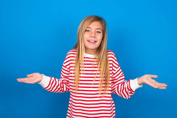 Wat dan nog? Portret van arrogante mooie blanke tiener meisje dragen gestreept shirt over blauwe studio achtergrond shrugging handen zijwaarts glimlachend hijgend onverschillig, vertellen iets voor de hand liggend. - Foto, afbeelding