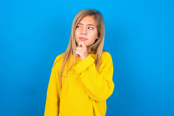 Przemyślana piękna blondynka nastolatka dziewczyna w żółtym swetrze nad niebieską ścianą trzyma podbródek i odwraca wzrok zamyślony wymyśla świetny plan - Zdjęcie, obraz