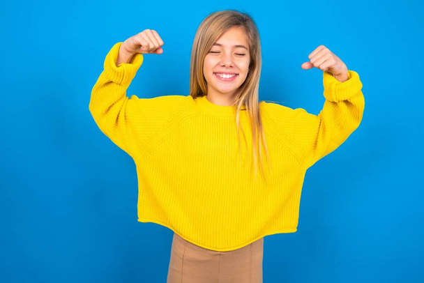 Forte potente bella ragazza bionda adolescente che indossa maglione giallo sopra la parete blu sorriso dentato, alza le braccia e mostra bicipiti. Guarda i miei muscoli.! - Foto, immagini
