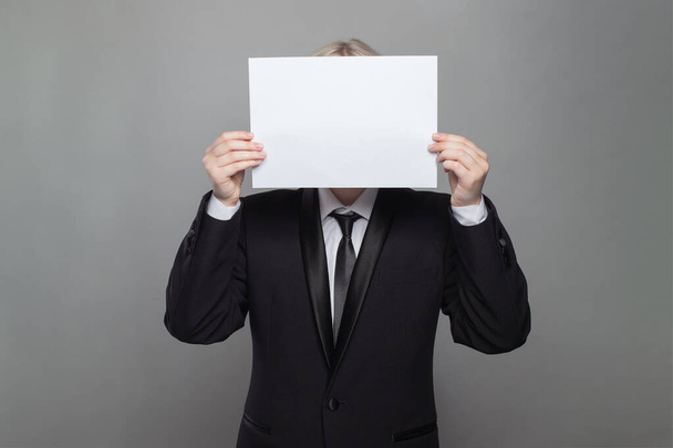 Ανώνυμος επιχειρηματίας κρύβει το κεφάλι του πίσω από κενό λευκό χαρτί σε γκρι φόντο στον τοίχο του στούντιο - Φωτογραφία, εικόνα
