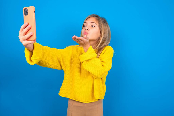 kaunis blondi teini tyttö yllään keltainen pusero yli sininen seinä puhaltaa ilmaa suudelma kameran älypuhelimen ja vie selfie, lähettää Mwah kautta online-puhelun. - Valokuva, kuva
