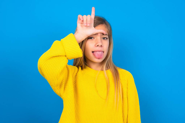 Funny beautiful blonde teen girl wearing yellow sweater over blue wall makes loser gesture mocking at someone sticks out tongue making grimace face. - Photo, Image