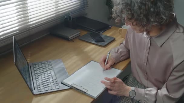Hochwinkelaufnahme einer reifen Frau mit grauem lockigem Haar, die als Psychologin am Schreibtisch in ihrem Büro sitzt und Behandlungspläne für ihre Patientin erstellt - Filmmaterial, Video