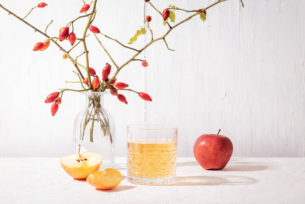 Frische reife Äpfel und ein Glas Apfelsaft oder Apfelmost auf weißem Tisch. Vase mit Hagebuttenzweigen im Hintergrund. Herbsternte, Erntedankkonzept. - Foto, Bild