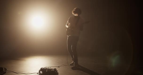 Full body handheld shot profesjonalnego milenijnego gitarzysty wykonującego muzykę rock and roll podczas poruszania się energicznie grając na gitarze elektrycznej w ciemnym pomieszczeniu przed żółtym reflektorem - Materiał filmowy, wideo