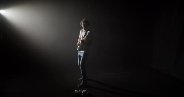 Přiblížit v pohodě moderní muž hraje na kytaru, zatímco stojí ve světle reflektorů na tmavém jevišti - Záběry, video
