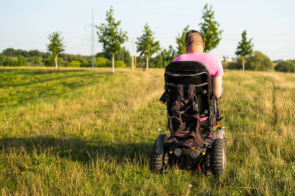 明るい日差しで照らされた緑色の牧草地でゆったりと散策している車椅子の男. 自動車事故後の足の怪我をした人は公園で自然を見ます - 写真・画像