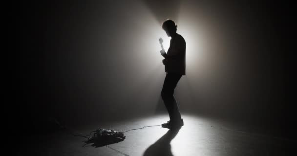 Cuerpo completo en tiempo real de guitarrista masculino enérgico parado en la oscuridad contra la luz brillante y tocando el instrumento musical eléctrico durante la actuación - Metraje, vídeo