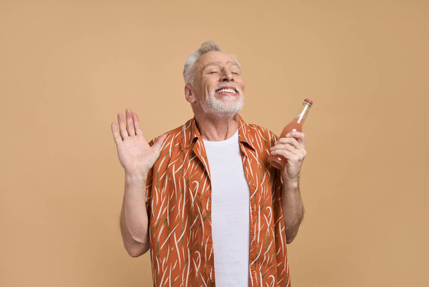 Kaukázusi idős férfi 60-69 éves, narancssárga ing, gazdaság frissítő limonádé, mosolygós, kifejező pozitív érzelmek, élvezi a nyári vakáció, pózol csukott szemmel elszigetelt bézs háttér - Fotó, kép