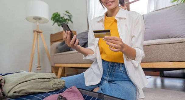 Mujer joven asiática usando teléfono inteligente reserva un hotel en línea con una tarjeta de crédito y preparar su equipaje para viajar. - Foto, imagen