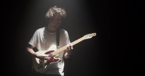 Prawdziwy czas tysiącletniego muzyka męskiego w luźnej koszuli grającego rock and rolla na gitarze elektrycznej podczas próby piosenki w ciemności z oświetleniem projektora - Materiał filmowy, wideo