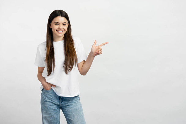 Beyaz tişört ve kot pantolon giyen gülümseyen genç kız fotokopi alanını işaret ediyor, mesaj için yer arıyor. Boş bir yere mutlu gençlik gösterisi, reklam - Fotoğraf, Görsel