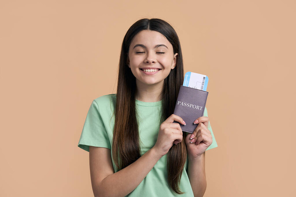 Caucasian happy pretty teenager traveler girl smiling cutely, holding boarding pass, waiting upcoming flight, enjoying happy summer holidays, posing with her eyes closed on beige isolated background. - Photo, Image