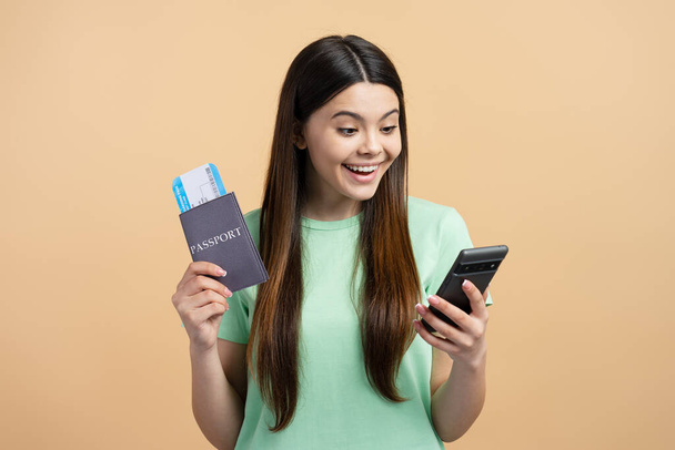 Portrait of cute smiling teenage girl holding mobile phone, passport with travel pass isolated on background - Photo, Image