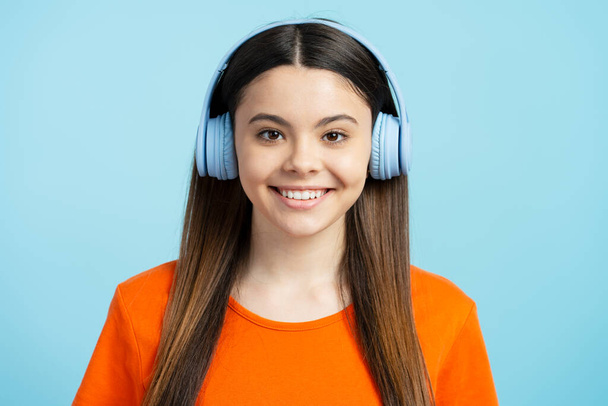 Attraktives lateinisches Teenagermädchen, das mit Kopfhörern Musik hört und isoliert auf blauem Hintergrund in die Kamera blickt. Glückliches Mädchen in stilvoller Freizeitkleidung, modernem Technologie-Konzept, genießen - Foto, Bild