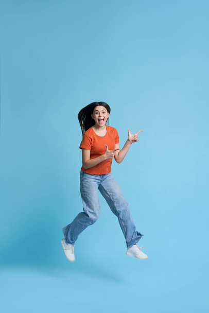 Прекрасна кавказька дівчина-підліток в помаранчевій футболці і блакитних джинсах, висловлюючи здивування і щастя, дивлячись на камеру, вказуючи на вільне рекламне простір, стрибаючи над синім ізольованим тлом - Фото, зображення