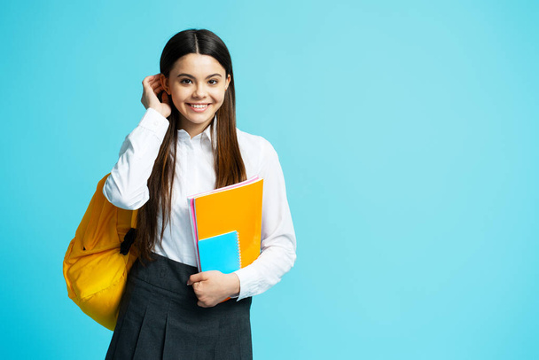 Portret van een positief tienermeisje in schooluniform, gele rugzak, kijkend naar de camera, haar gefixeerd op blauwe achtergrond. Terug naar school - Foto, afbeelding