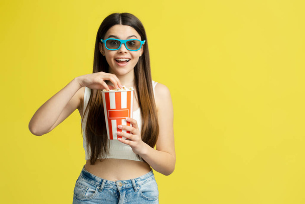 Muchacha emocionada sosteniendo cubo con palomitas de maíz viendo película en gafas 3d aisladas sobre fondo amarillo - Foto, imagen