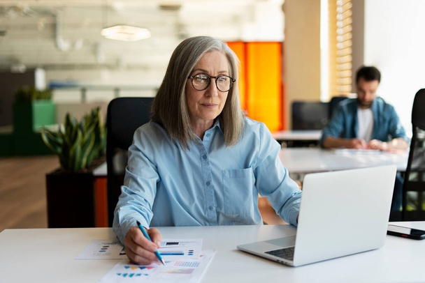 Серйозна зріла бізнес-леді використовує ноутбук, роблячи нотатки, працюючи над проектом в сучасному офісі. 60 років жінка, фінансистка, що працює з документами на робочому місці. Успішний бізнес, кар'єра  - Фото, зображення
