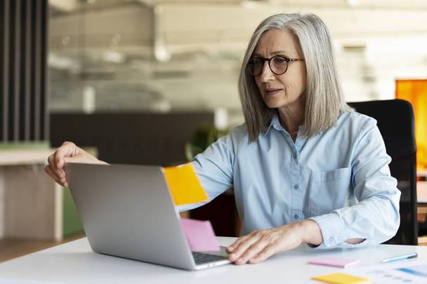 Attractive gray haired senior business woman wearing eyeglasses using laptop computer, working online sitting in modern office, Successful manager using sticky notes, agile methodology. Scrum concept - Photo, Image