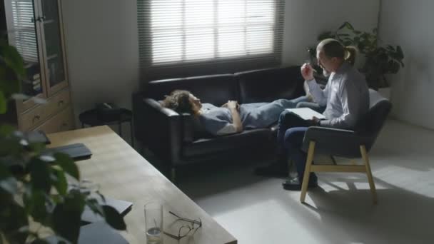 Vysoký úhel dlouhý záběr mladé ženy s post-traumatickou poruchou ležící na trenéra mluvit s psychoterapeutem během sezení - Záběry, video