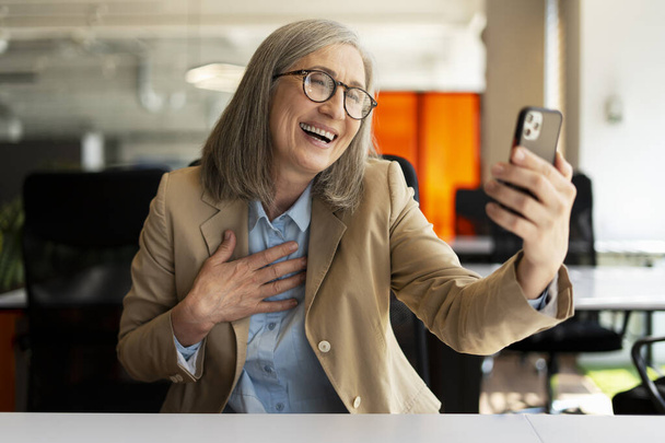 Portret szczęśliwej siwowłosej starszej kobiety trzymającej telefon komórkowy, komunikacja online w nowoczesnym biurze. Koncepcja połączenia wideo - Zdjęcie, obraz