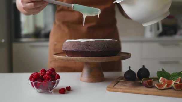 Kaunis tyytyväinen naispuolinen kondiittori valmistaa kakun, jossa on valkoista kermaa ja sisustettuja marjoja viihtyisässä keittiössä. ruoka-, leivonta- ja ruoanlaittokonsepti  - Materiaali, video