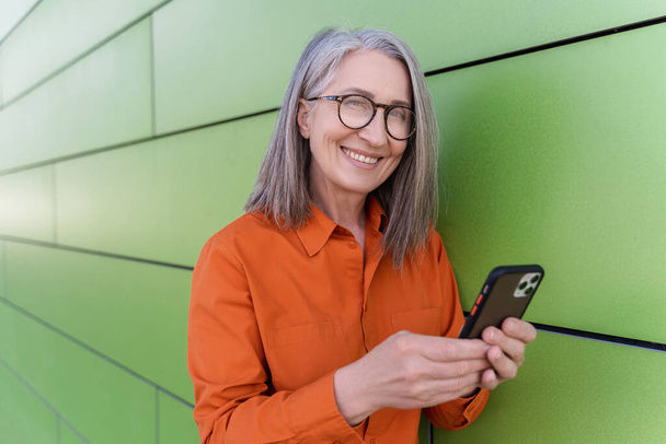 Portrait smiling gray haired senior woman holding smartphone using mobile app shopping online standing on the street. Happy 60 years old female in stylish eyeglasses reading text message. Technology  - Fotó, kép