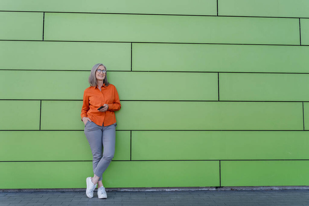 Plná délka portrét módní stylové krásné zralá šedovlasá žena 60 let, v oranžové košili a modré džíny, stojí u zelené kovové stěny, s mobilním telefonem v ruce - Fotografie, Obrázek