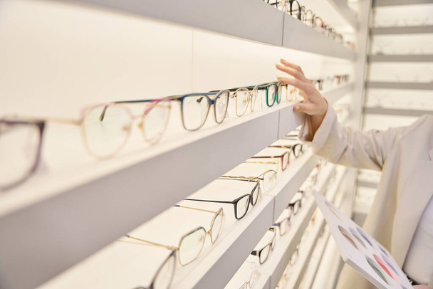 Woman consultant selects the right frames on the display window, in the salon there is a wide selection of glasses - Photo, Image